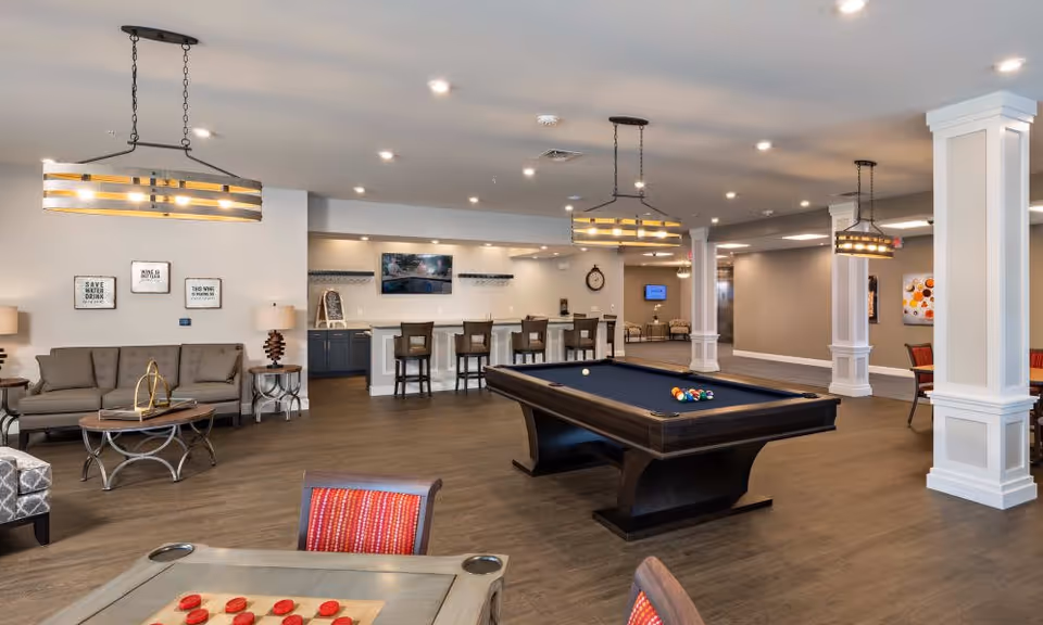 A spacious common area in a senior living facility featuring a pool table with balls arranged for a game, a checkers table in the foreground, a seating area with a sofa and armchairs, a bar counter with stools, and modern pendant lighting fixtures hanging from the ceiling. The room has wood flooring, neutral-colored walls, and decorative wall art.