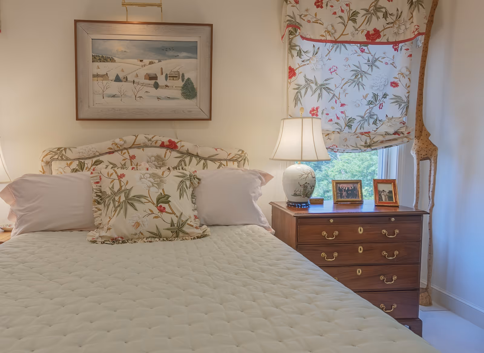 A cozy bedroom with a bed featuring a floral patterned headboard and matching pillow. There are two white pillows on the bed. To the right of the bed is a wooden nightstand with a decorative lamp, two framed photos, and a window with a floral curtain. A tall wooden giraffe sculpture stands in the corner near the window. A framed painting of a winter landscape hangs on the wall above the bed.