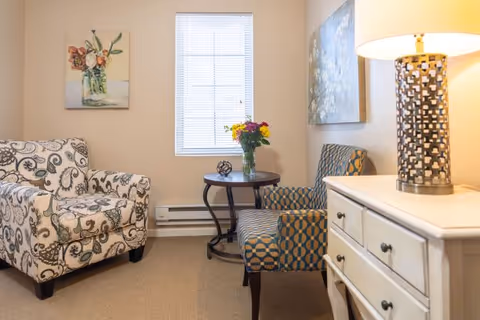A cozy sitting area with two patterned armchairs, a small round wooden table with a vase of colorful flowers, a white chest of drawers with a decorative lamp, and a window with blinds letting in natural light. The walls are decorated with floral paintings.