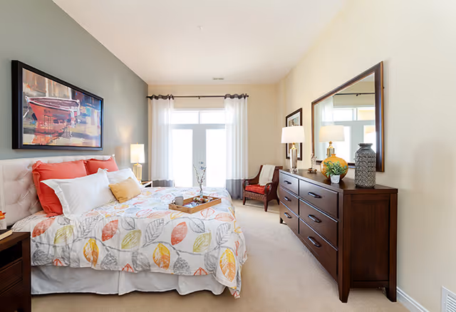 A bright and cozy bedroom with a large bed featuring a colorful leaf-patterned bedspread and multiple pillows. There is a wooden nightstand with a lamp on one side of the bed, and a large wooden dresser with a mirror, decorative items, and a lamp on the opposite wall. A comfortable chair is placed near a window with sheer white curtains allowing natural light to fill the room. A framed artwork hangs above the bed.