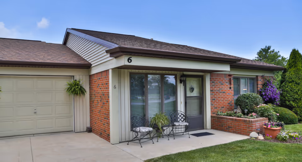 Exterior view of a single-story brick and siding residential building with a garage, a small patio with two chairs and a table, potted plants, and a well-maintained garden with shrubs and flowers under a clear blue sky.
