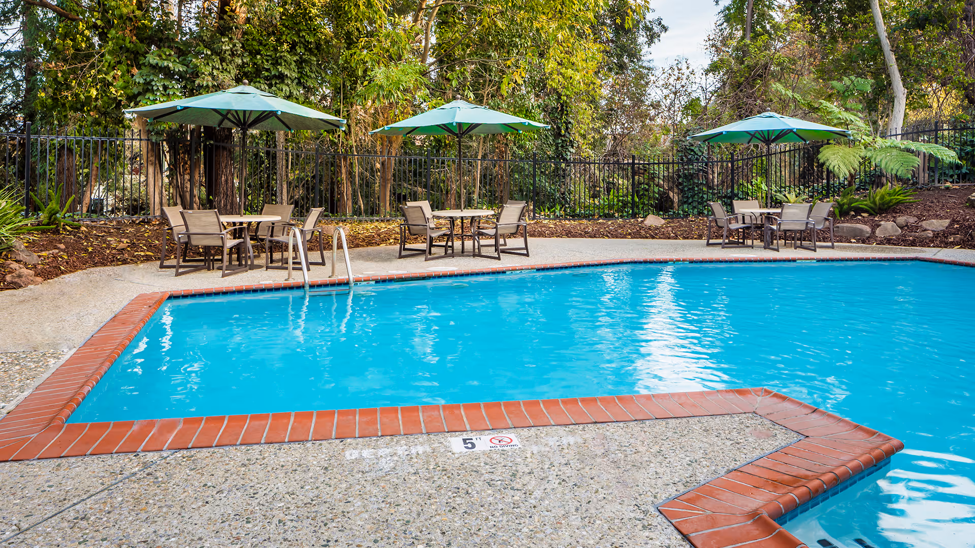 Outdoor swimming pool with clear blue water surrounded by a concrete deck with red brick edging. There are three patio tables with green umbrellas and chairs around the pool. The area is enclosed by a black metal fence and surrounded by trees and greenery.