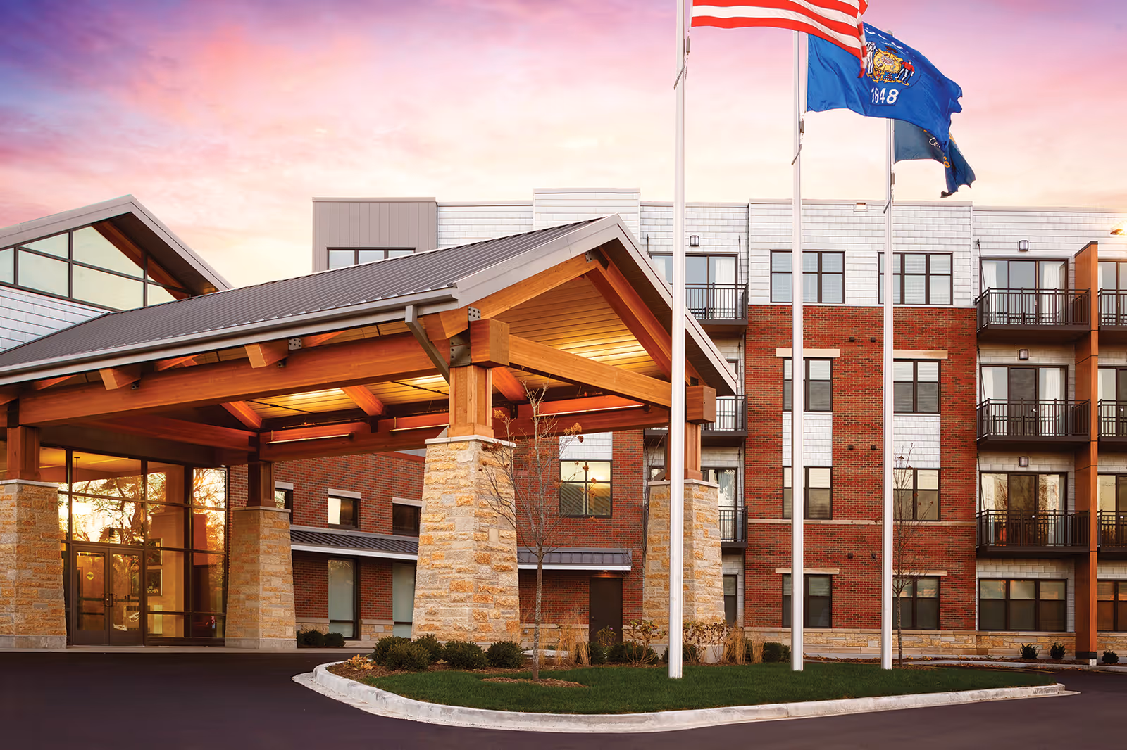 Exterior view of a senior living facility named Ciel of Shorewood during sunset, featuring a covered entrance with wooden beams and stone pillars, three flagpoles with flags, and a multi-story building with balconies and large windows.