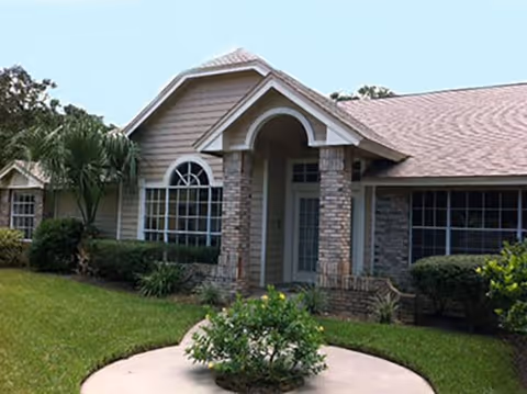 Front exterior view of a single-story residential building with a gabled roof, stone and siding facade, large windows, and a small landscaped area with bushes and a circular concrete path in front.