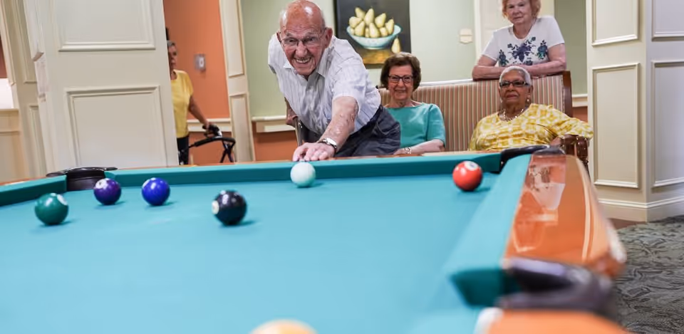 An elderly man is playing pool, aiming to hit the cue ball on a billiards table. Three elderly women are seated and standing nearby, watching him play in a well-lit room with light-colored walls and a painting of pears on the wall.
