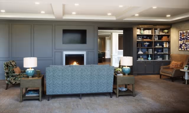 A cozy living room with a blue patterned sofa facing a fireplace set in a gray paneled wall. Two wooden side tables with blue lamps flank the sofa. To the right, there is a built-in dark wood bookshelf filled with decorative items and books, and a brown armchair with a cushion next to a small round table. The room has recessed ceiling lights and a beige carpeted floor.