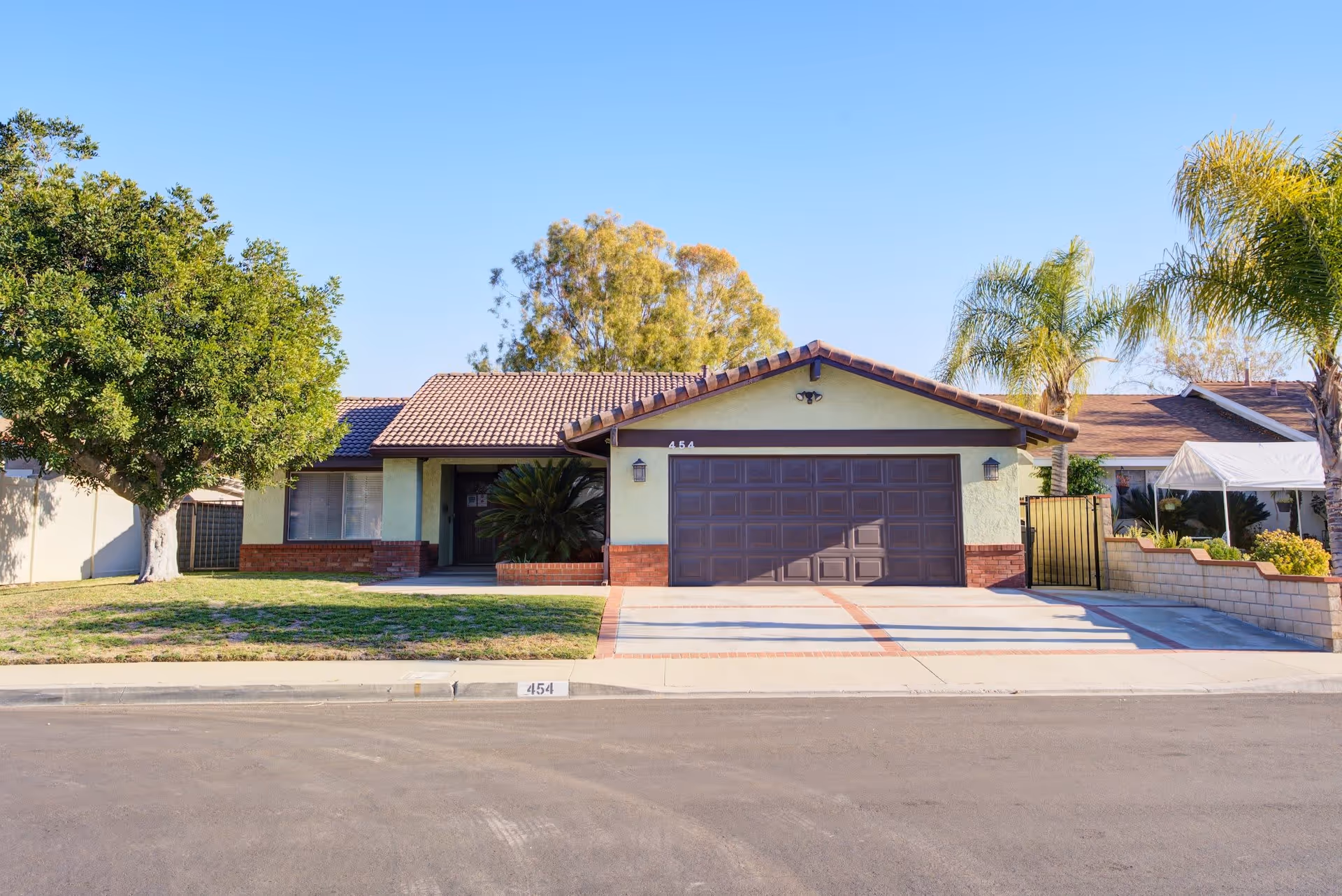 Front exterior view of a single-story house with a tiled roof, a large garage door, a driveway, a green lawn with a tree on the left, and palm trees on the right under a clear blue sky.