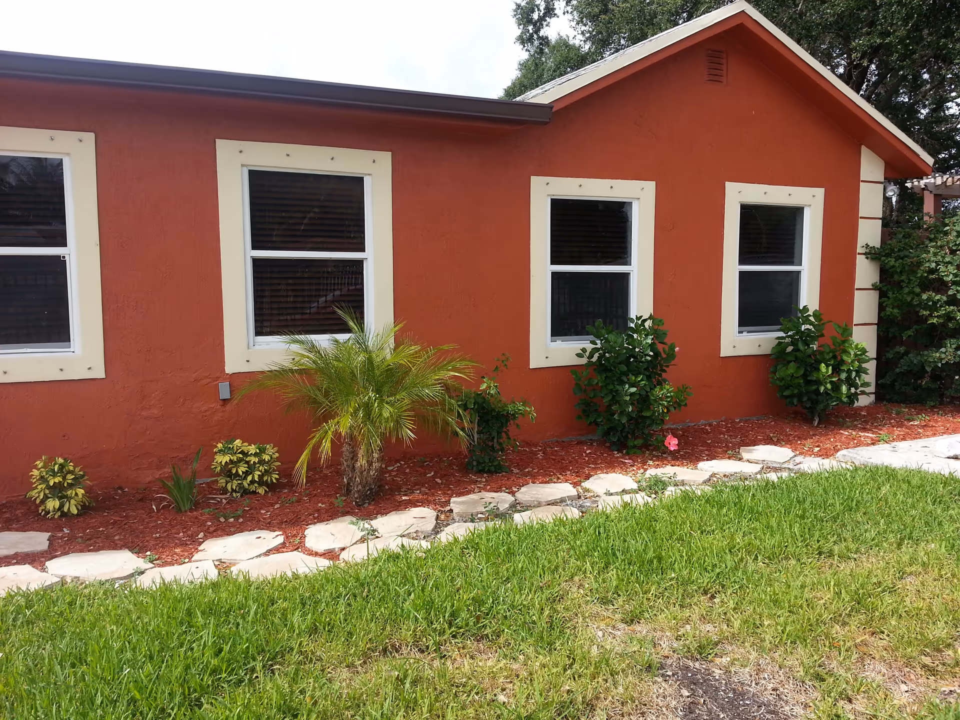 Exterior view of a red building with four windows framed in white. In front of the building, there is a garden bed with small bushes, a palm plant, and red mulch, bordered by irregularly shaped stepping stones and green grass.