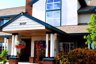 Entrance of a senior living facility with a covered porch supported by white columns and brick bases. The building has a large window above the entrance and hanging flower baskets on either side of the doorway. There are shrubs and greenery around the entrance area.