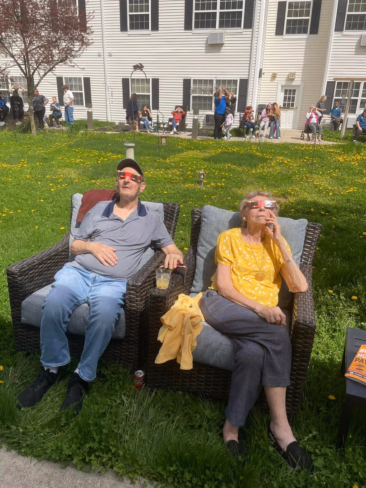 Two elderly people sitting outdoors on cushioned wicker chairs on a grassy lawn, wearing special glasses and looking up at the sky. Behind them is a white multi-story building with several windows and people standing or sitting along a paved walkway.