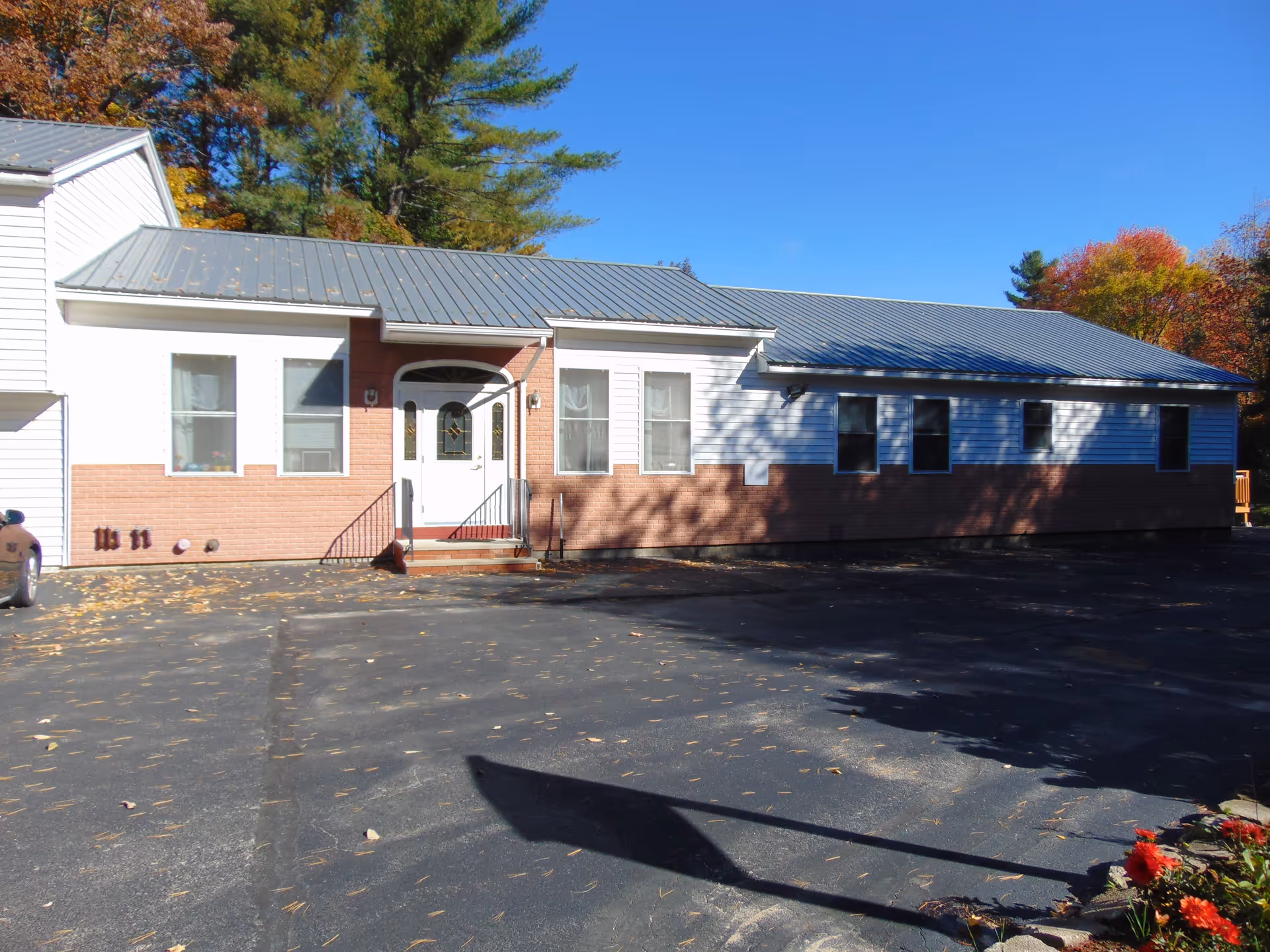 Exterior view of a single-story building with white siding and a brick lower half, featuring a metal roof and a white front door with glass panels. The building is surrounded by a paved parking area with some fallen leaves and trees with autumn foliage in the background under a clear blue sky.