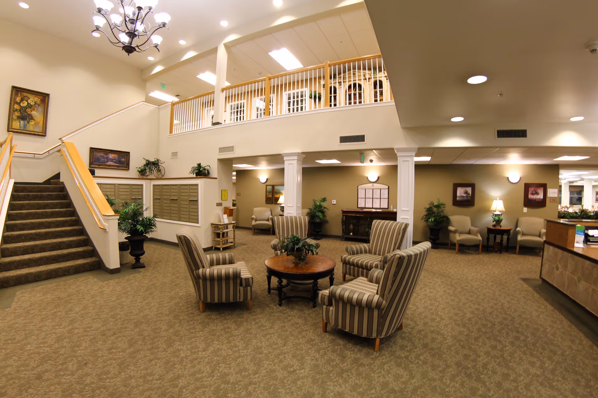 Interior view of a senior living facility lobby with a seating area consisting of four striped armchairs around a round wooden table with a plant centerpiece. There is a staircase on the left side leading to an upper floor with white railings. The walls are decorated with framed paintings and there are several potted plants around the room. The lighting includes ceiling lights and a chandelier above the staircase.
