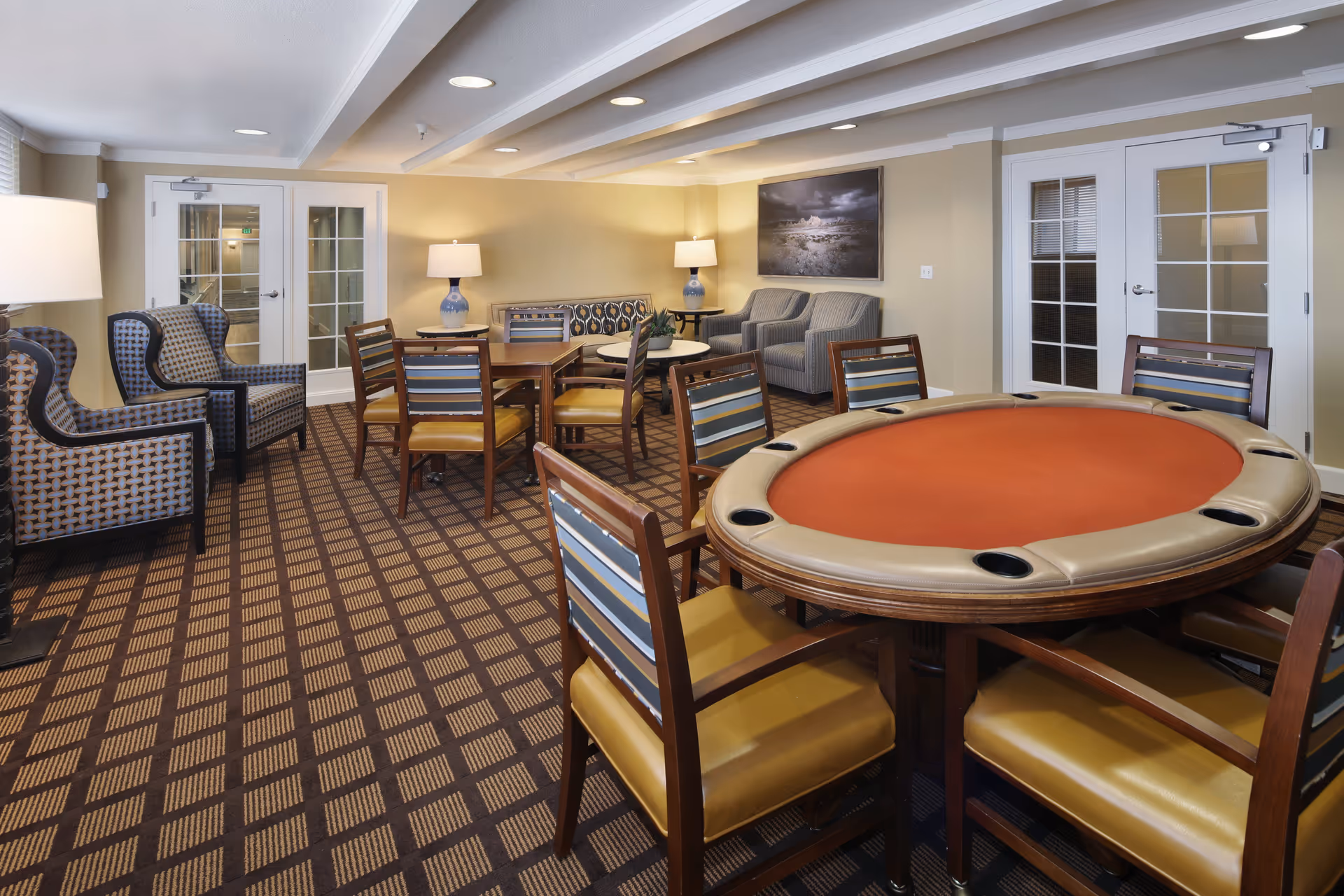 A cozy common area in a senior living facility featuring a round poker table with padded edges and six chairs, a square wooden table with four chairs, two patterned armchairs, a sofa, two armchairs, two table lamps, and a framed landscape photograph on the wall. The room has a checkered carpet and beige walls with white trim.