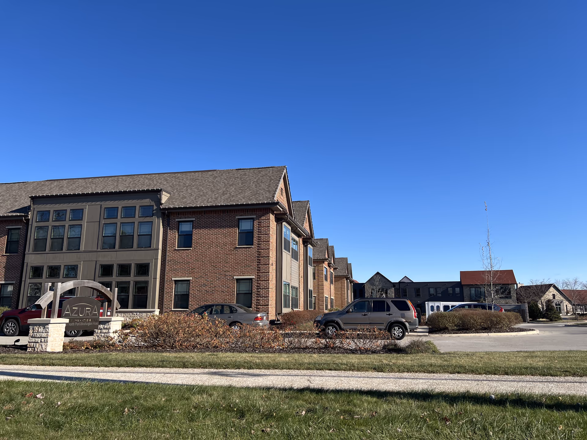 Exterior view of Azura Assisted Living and Memory Care of Fox Point building under a clear blue sky, with parked cars and a sign in front of the building.