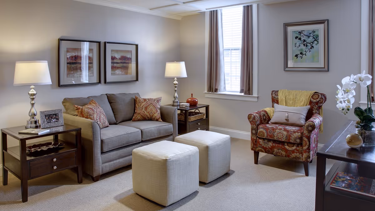 A cozy living room with a gray sofa adorned with patterned cushions, two beige ottomans in front, a floral-patterned armchair with a yellow throw and a pillow, two side tables with lamps, framed artwork on the walls, a window with curtains, and a table with a white orchid plant and decorative items.