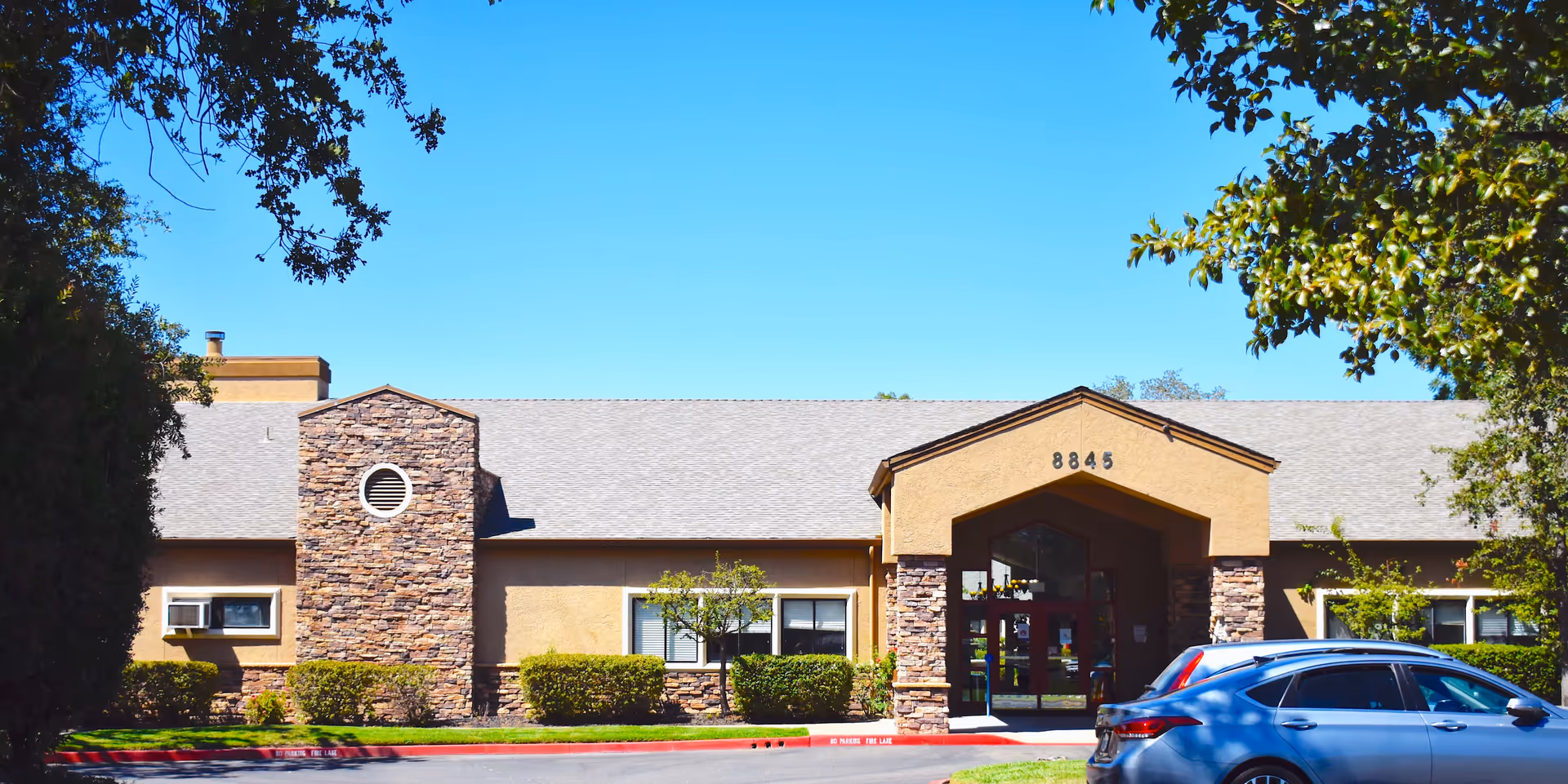 Front exterior view of a single-story building with stone and beige stucco facade, a peaked entrance with the number 8845 above it, surrounded by trimmed bushes and trees under a clear blue sky. A blue car is parked in front of the building.