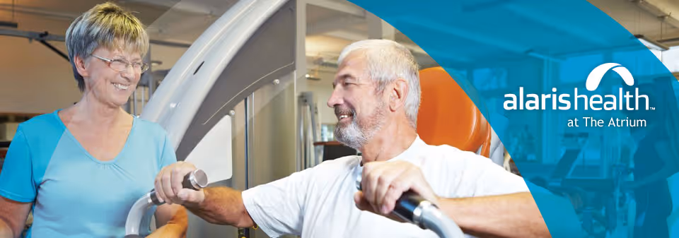 An elderly man using exercise equipment in a fitness or rehabilitation setting, assisted by a smiling female caregiver or therapist. The background shows gym equipment and a bright, welcoming environment.