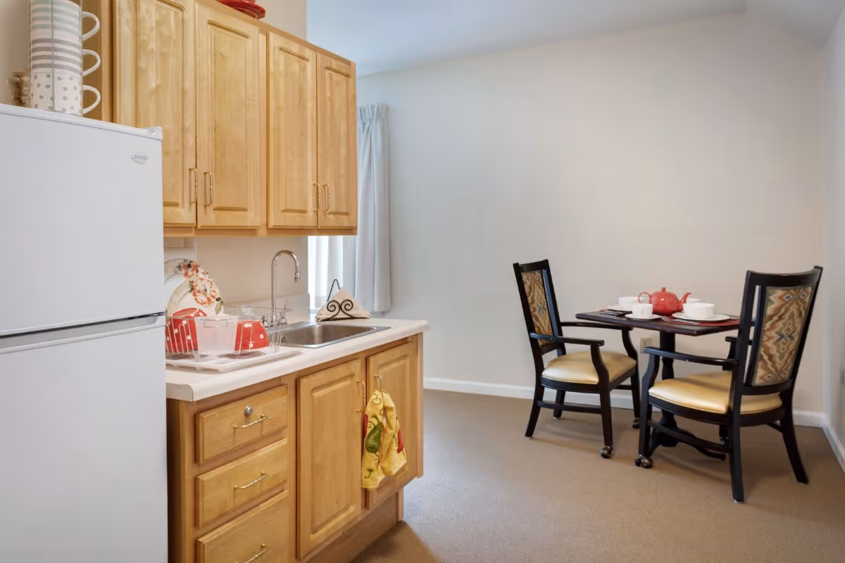 A small kitchen area with light wooden cabinets, a white refrigerator, a sink, and a countertop with dishware and a drying rack. Adjacent to the kitchen is a small dining area with a dark wooden table set for tea and two upholstered chairs with patterned backs and beige seats. The walls are plain and light-colored, and the floor is carpeted.