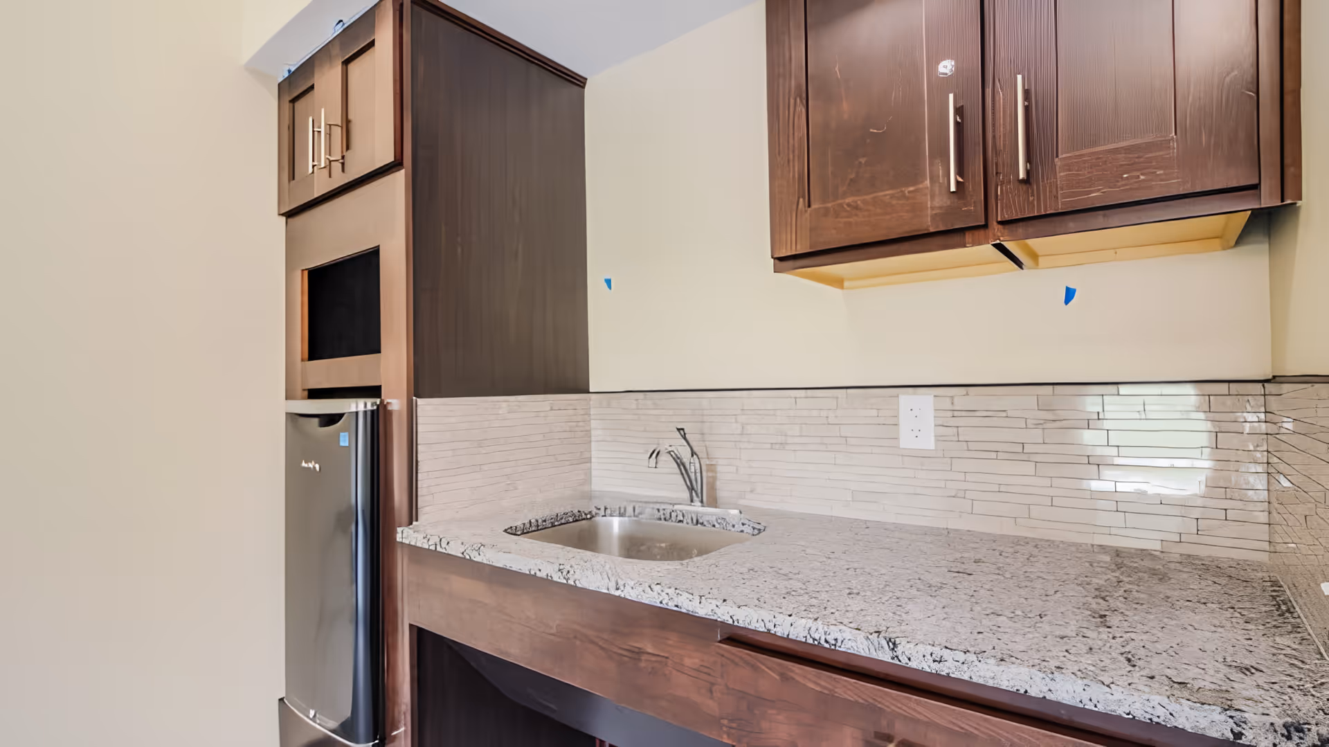 A modern kitchen area with dark wood cabinets, a small stainless steel sink, a granite countertop, and a compact refrigerator. The backsplash features horizontal beige tiles, and there is an electrical outlet on the wall above the countertop.