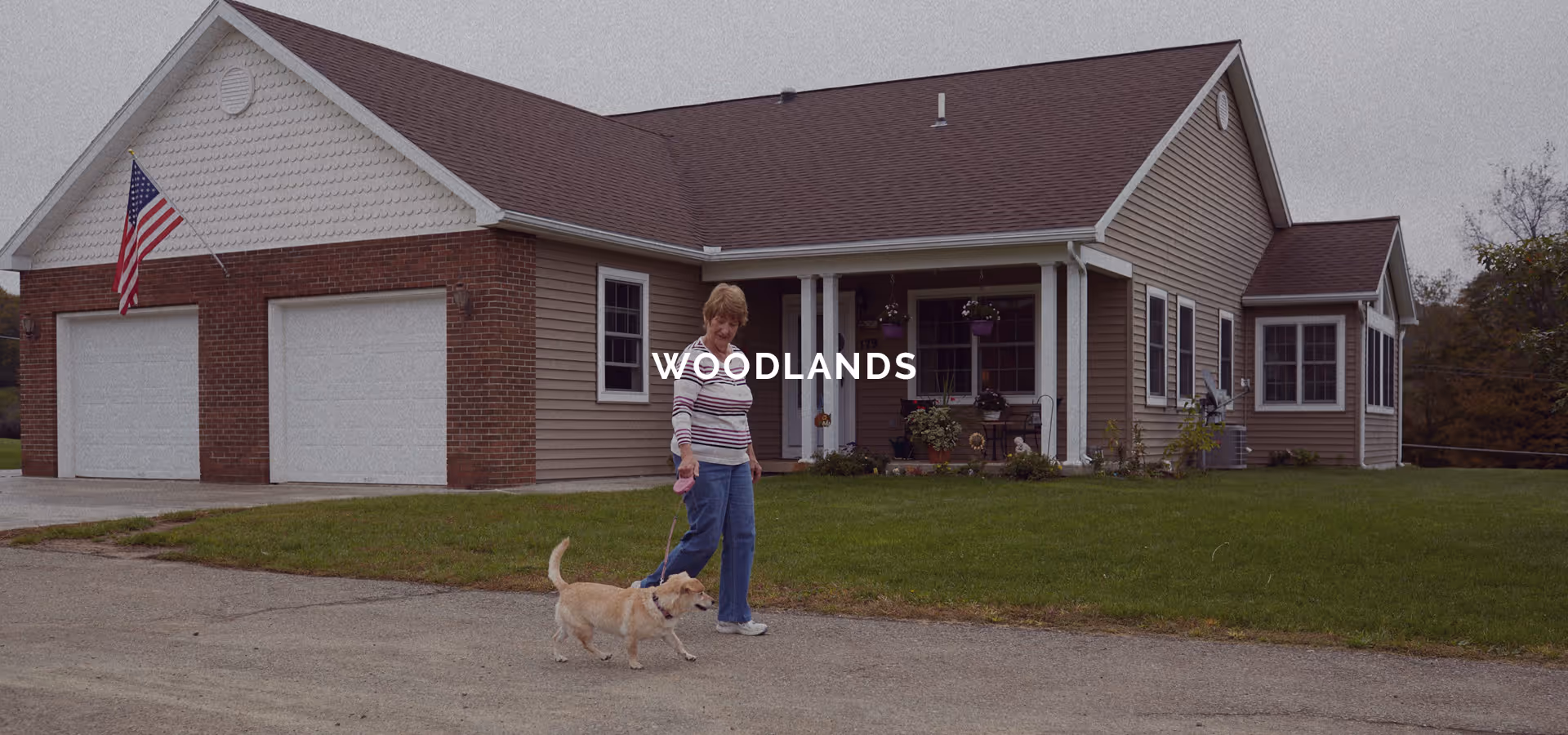 A woman walking a small dog on a leash in front of a single-story house with a two-car garage. The house has beige siding, a brick facade on the garage, an American flag mounted near the garage, and a well-maintained lawn.