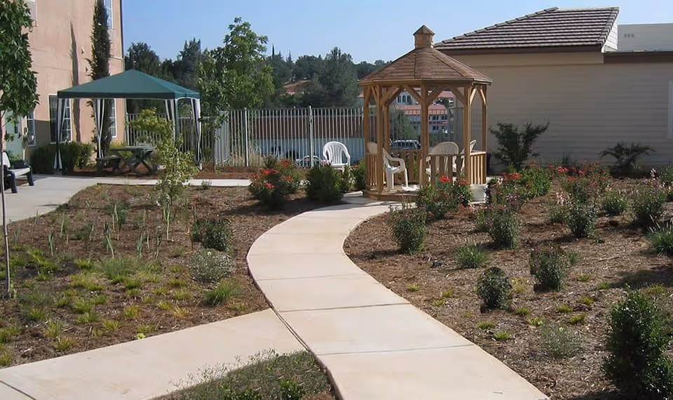 A paved walkway curves through a garden area with small bushes and plants. To the right, there is a wooden gazebo with white plastic chairs inside. On the left side, there is a green canopy covering a picnic table. In the background, there is a building and a metal fence with trees and hills beyond.