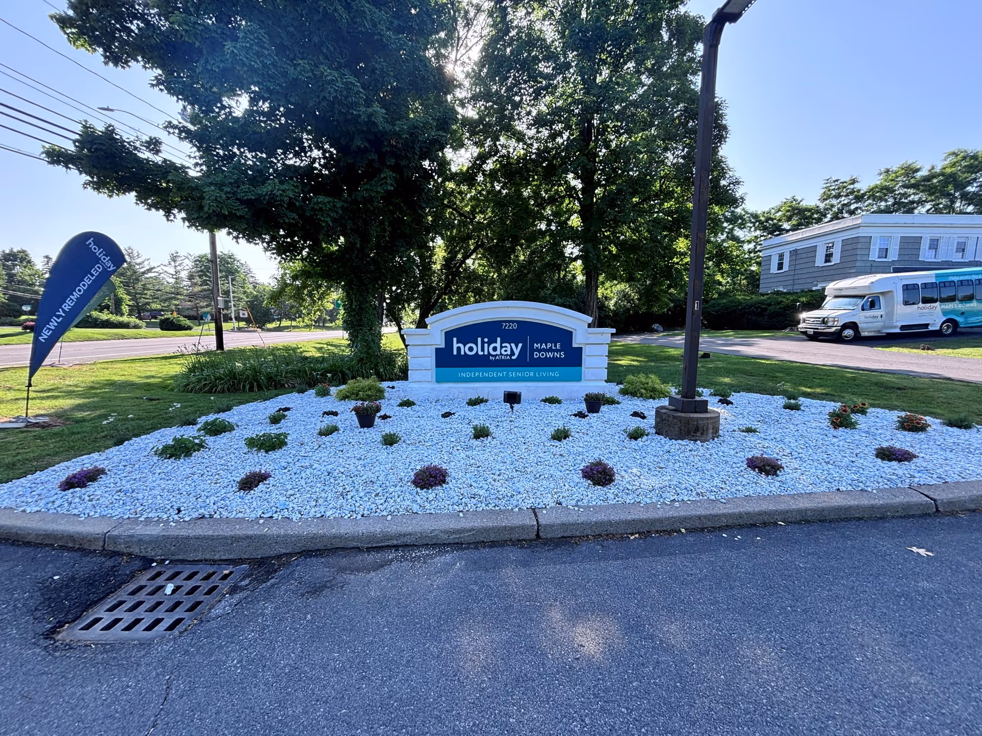 Outdoor view of the entrance sign for Holiday Maple Downs independent senior living facility, surrounded by white decorative rocks and small plants, with a flag sign on the left that reads 'NEWLY REMODELED' and a shuttle bus parked on the right side near a building.