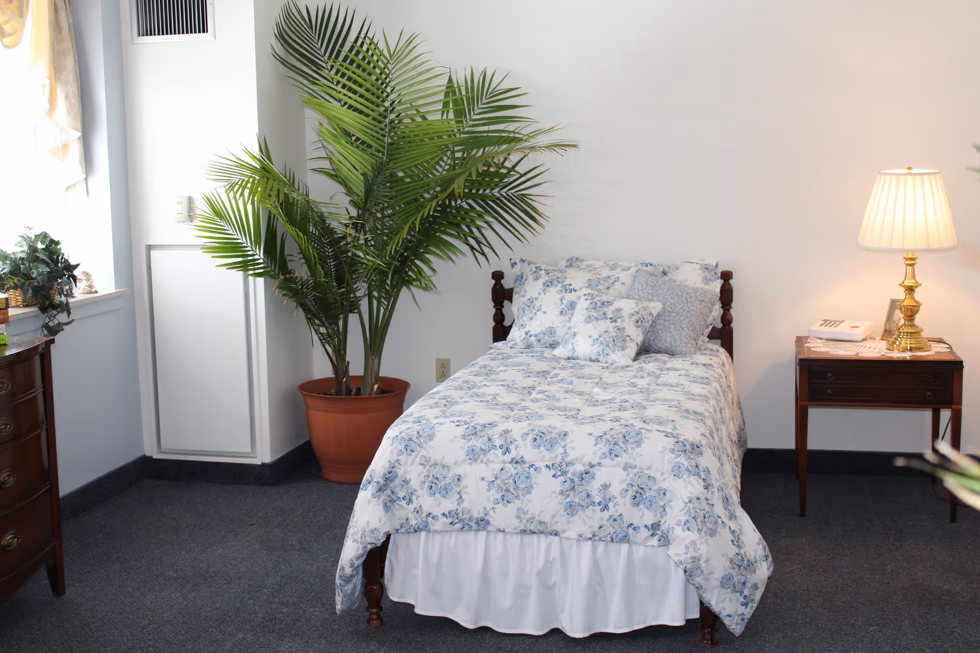 A neatly made single bed with a floral blue and white comforter and matching pillows in a room. To the right of the bed is a wooden nightstand with a brass lamp and a telephone. To the left of the bed is a large potted plant. The room has a window with sheer curtains and a wooden dresser partially visible on the left side.