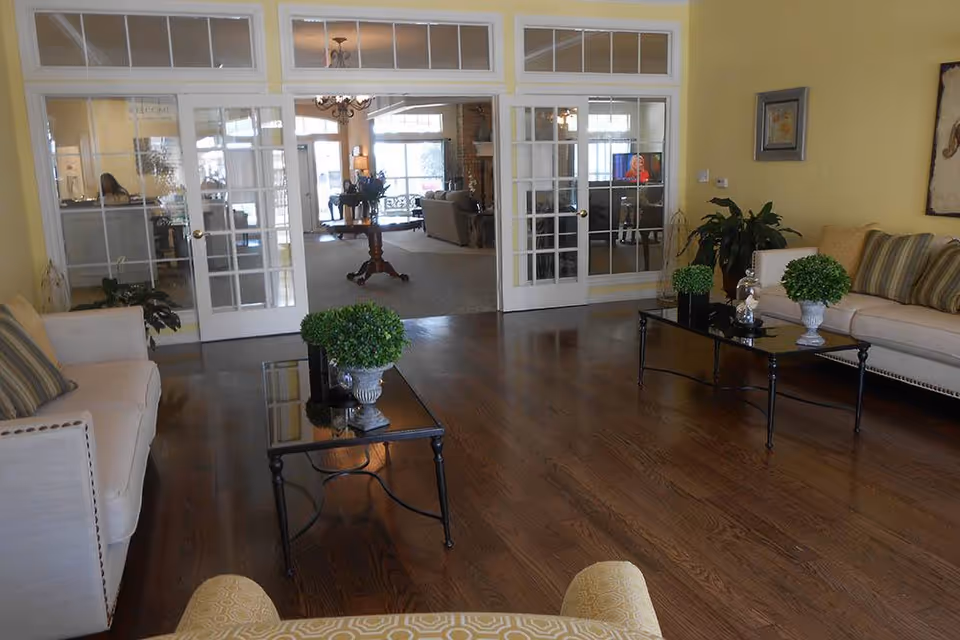 A bright and spacious living room with wooden flooring, two beige sofas with striped cushions, two glass coffee tables with green potted plants, and large glass-paneled double doors leading to another room with more seating and a table with flowers.