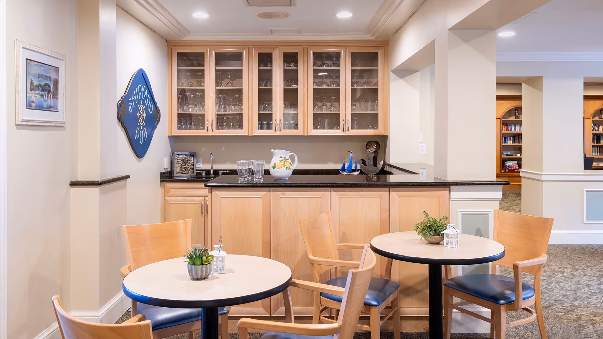 A small dining area in a senior living facility with two round tables, each surrounded by wooden chairs with blue cushions. Behind the tables is a wooden cabinet with glass doors displaying glassware, a countertop with a pitcher and glasses, and nautical-themed decorations on the wall. The space is well-lit and has a cozy, welcoming atmosphere.