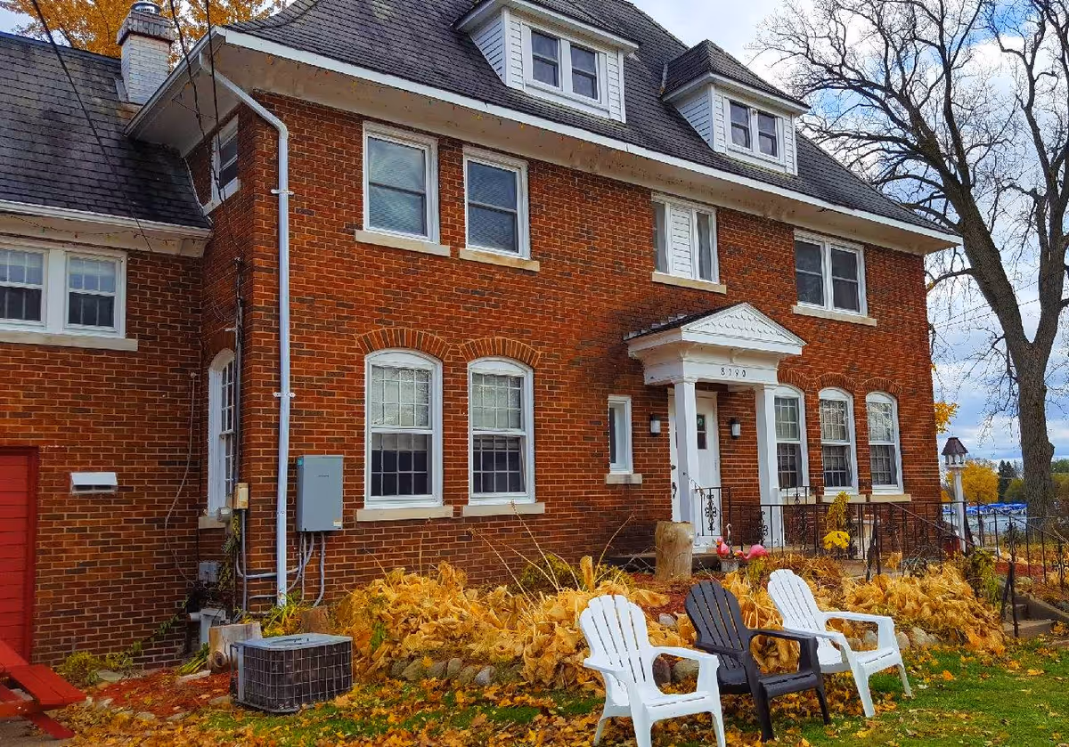 Exterior view of a two-story brick building with white-framed windows and a small porch with white columns. In front of the building, there are three plastic chairs, two white and one black, placed on a lawn with fallen autumn leaves. Trees with bare branches and some yellow foliage are visible around the building under a partly cloudy sky.