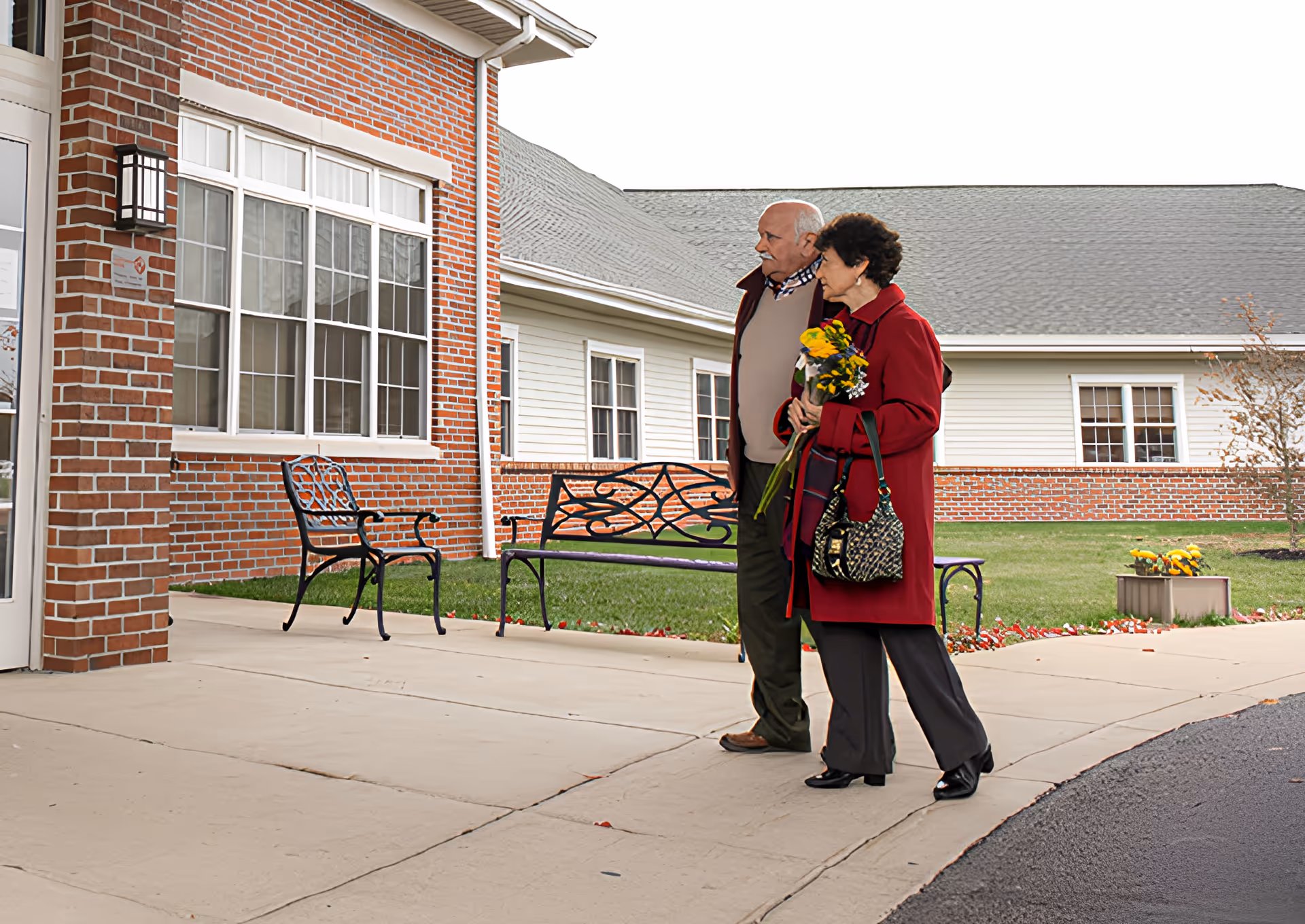 An elderly couple walking arm in arm outside a brick and siding building. The woman is holding a bouquet of flowers and carrying a patterned handbag. There are metal benches and a small flower bed near the building entrance.