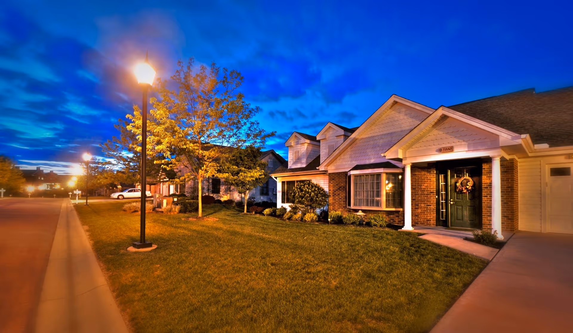 A well-lit residential street at dusk with single-story houses featuring front porches, manicured lawns, and trees. Street lamps illuminate the sidewalk and road, creating a warm and inviting atmosphere under a deep blue sky.