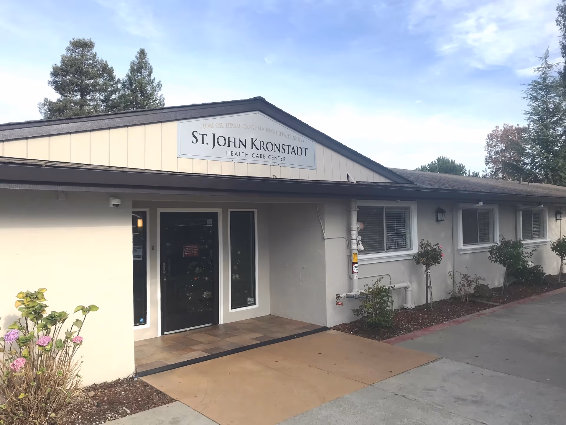 Exterior front view of St. John Kronstadt Health Care Center building with a sign above the entrance door, surrounded by small bushes and trees under a partly cloudy sky.