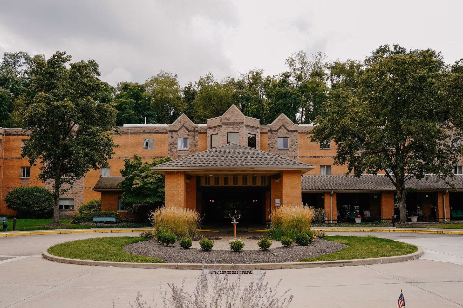 Front exterior view of a senior living facility named Asbury Heights, featuring a three-story brick building with a covered entrance, surrounded by trees and landscaped greenery.