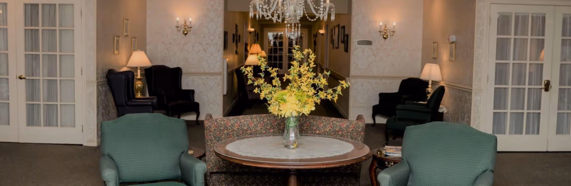 A cozy senior living community lounge area with two green armchairs and a floral-patterned loveseat arranged around a round wooden table with a lace doily and a vase of yellow flowers. The background features a hallway with wall sconces, table lamps, and framed pictures, along with French doors on either side.
