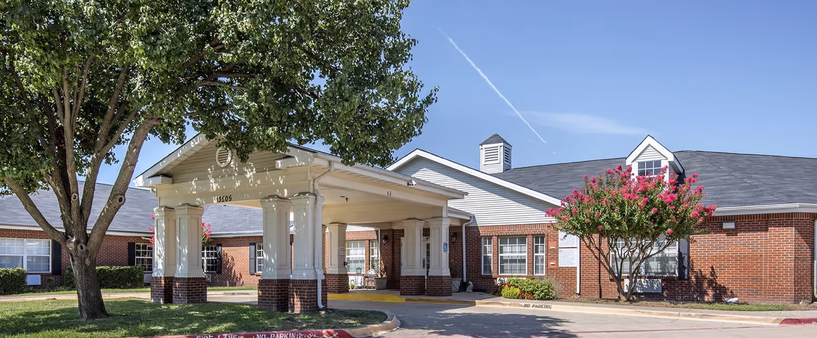 Exterior view of a single-story brick building with a covered entrance supported by white columns. There are trees and landscaping around the building under a clear blue sky.