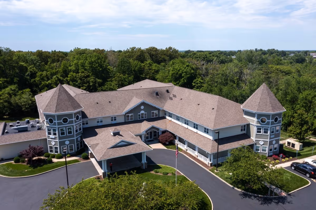 Aerial view of Cedar Creek Of Shiloh, a large senior living facility with two prominent turret-style towers, surrounded by trees and greenery. The building has a covered entrance driveway and a circular driveway with landscaped areas and a flagpole in front.