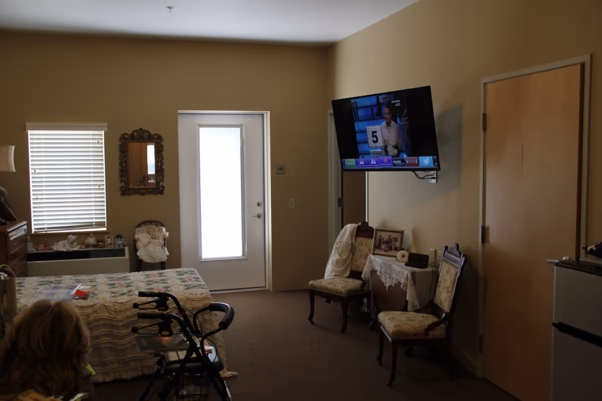 A senior living facility bedroom with a bed covered in a patterned quilt, a walker in front of the bed, two upholstered chairs, a small table with a lace cloth and framed pictures, a wall-mounted flat screen TV, a window with blinds, and a door with a frosted glass panel.