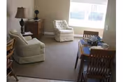 A cozy living room area with two beige armchairs, a wooden side table with a lamp and decorative item, and a dining table with four wooden chairs. A large window lets in natural light, brightening the neutral-toned room.