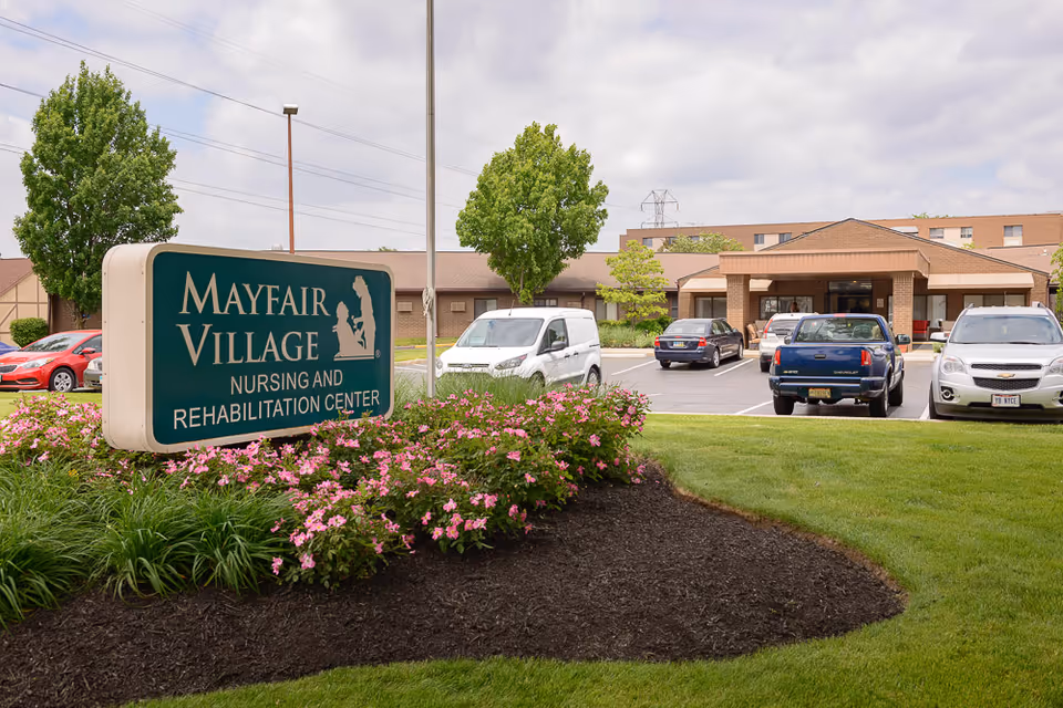Exterior view of Mayfair Village Nursing and Rehabilitation Center showing the building entrance, parking lot with several parked cars, a green lawn, and a sign with the facility's name surrounded by pink flowers and greenery.