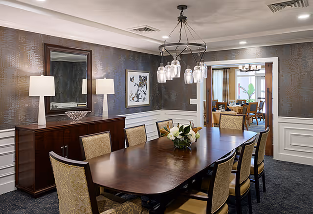 A formal dining room with a long wooden table surrounded by eight upholstered chairs. The room features a sideboard with two table lamps and a decorative bowl, a large mirror above the sideboard, and a framed artwork on the wall. A chandelier with multiple glass shades hangs above the table. Through an open doorway, another room with additional seating and a chandelier is visible.