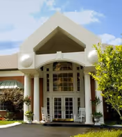 Front exterior view of McKnight Place Assisted Living & Memory Care building featuring a large entrance with tall white columns, a peaked roof overhang, glass double doors, and two white rocking chairs on the porch. There are plants and trees around the entrance and a clear blue sky above.