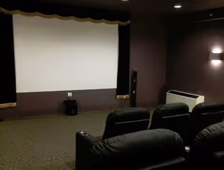 A small theater room with a large white projection screen on the front wall, several black leather recliner chairs arranged in rows facing the screen, dim lighting, and a carpeted floor.