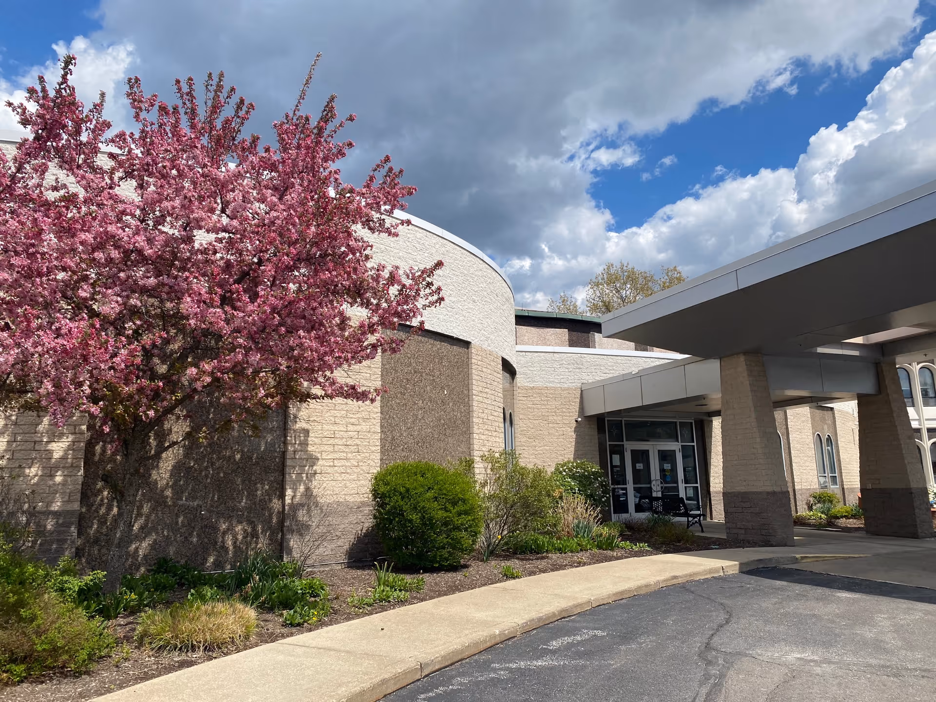 Exterior view of Light of Hearts Villa building with a covered entrance, beige brick walls, and a blooming pink tree in the landscaped garden under a partly cloudy sky.