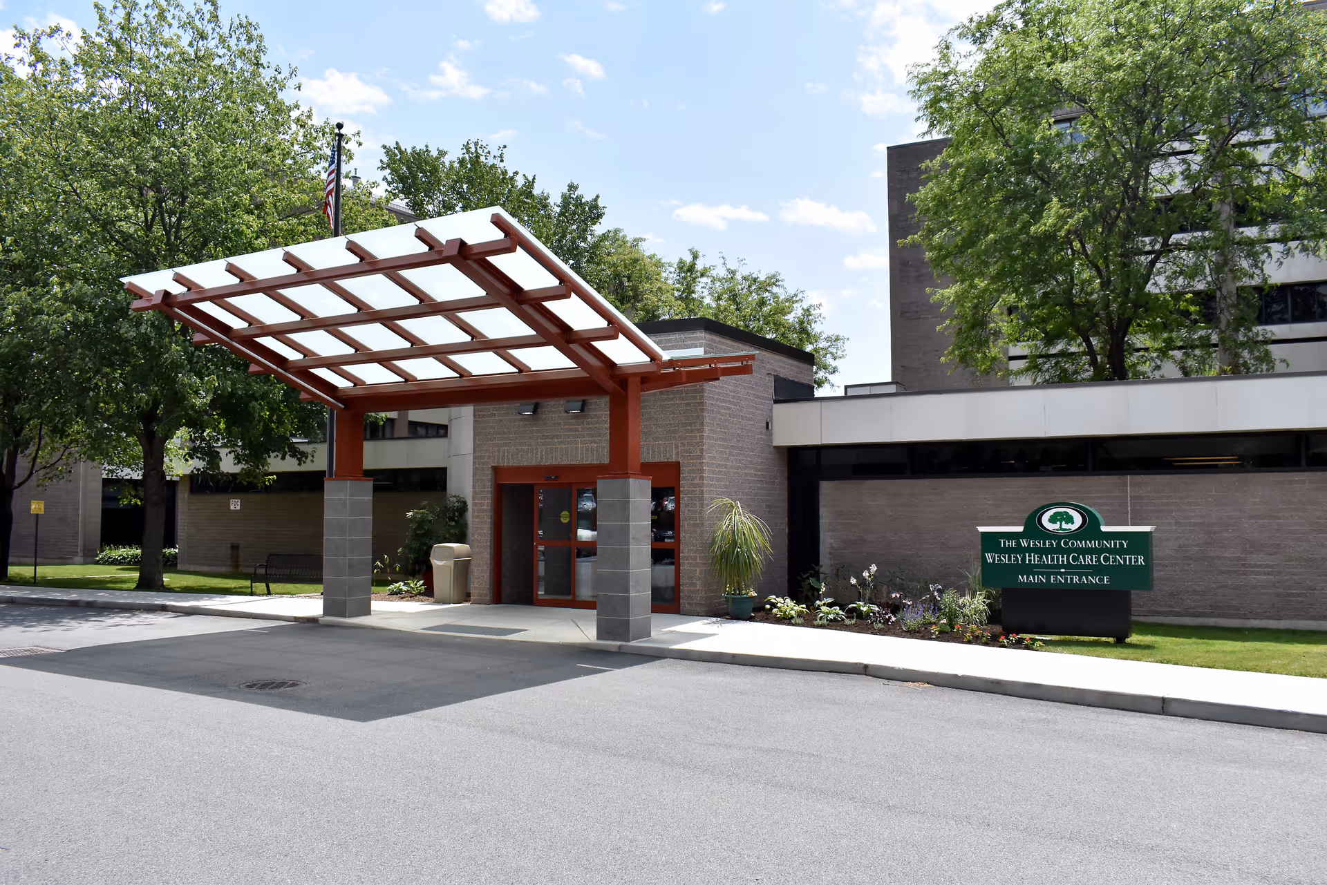 Exterior view of The Wesley Community - Wesley Health Care Center main entrance with a covered drop-off area, surrounded by trees and landscaping under a partly cloudy sky.