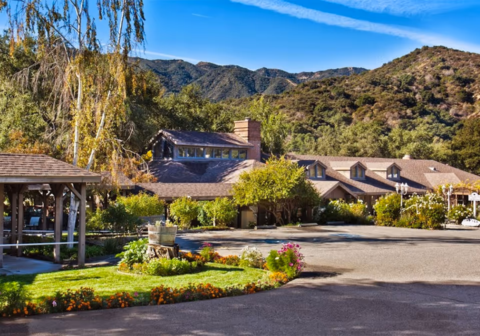 Exterior view of a senior living facility named Twelve Oaks surrounded by greenery and mountains in the background. The building has a brown roof with multiple dormer windows and a chimney. There is a gazebo on the left side and a landscaped garden with flowers and shrubs in the foreground.