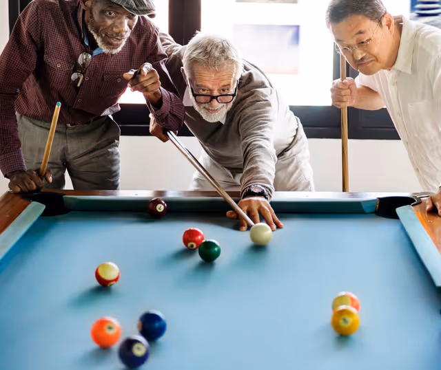 Three elderly men playing pool together indoors, one man is aiming with a cue stick while the other two watch closely.