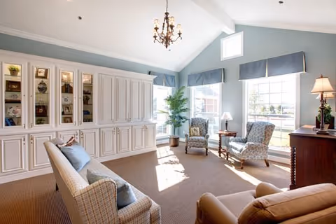A bright and spacious living room with large windows letting in natural light. The room features a chandelier, built-in white cabinetry with glass display shelves, two patterned armchairs with a small table between them, a sofa with cushions, a brown recliner, a floor lamp, and a potted plant near the windows.