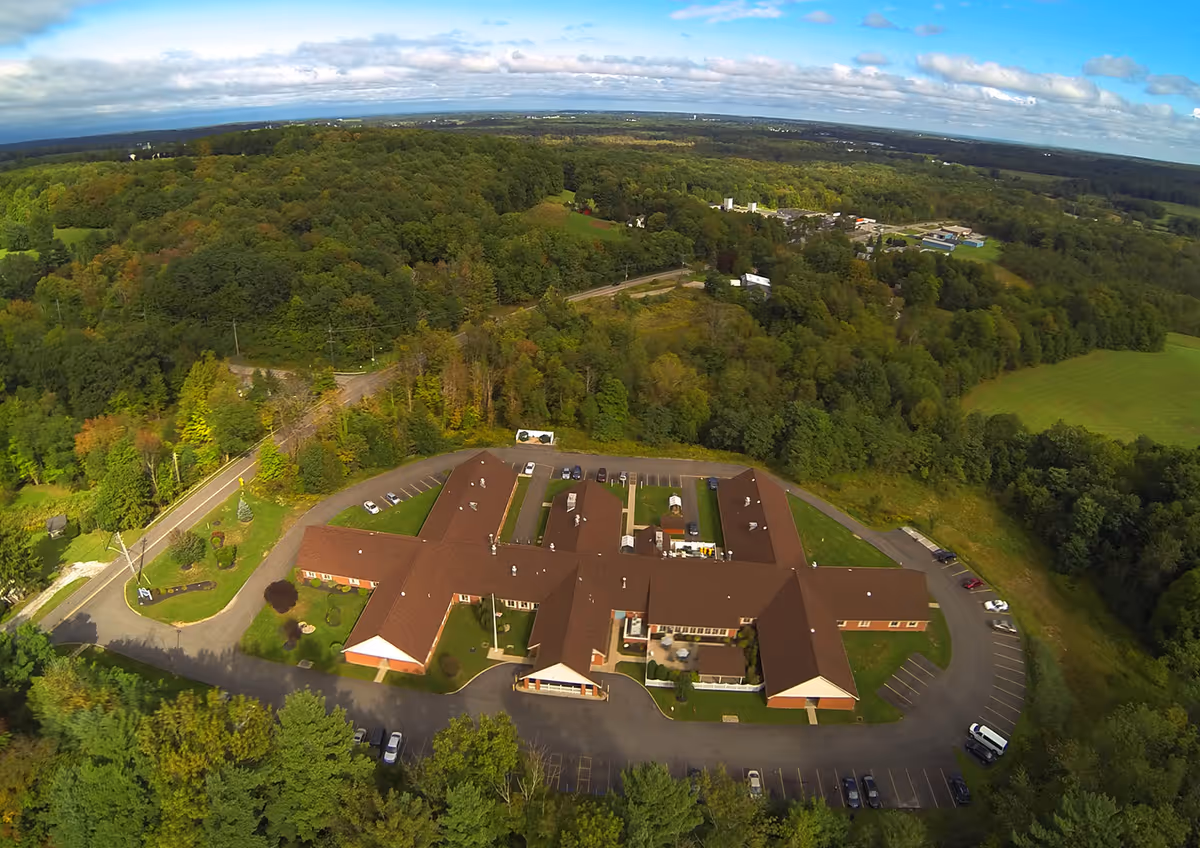 Aerial view of Burton Health Care Center surrounded by dense green trees and open fields under a partly cloudy blue sky. The building has a brown roof and is shaped in a U-formation with parking lots around it.