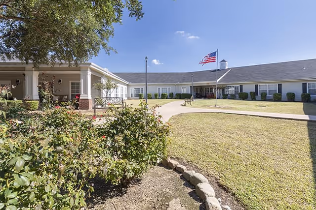 Outdoor courtyard area of a senior living facility with a well-maintained lawn, bushes, and a large American flag on a flagpole. The building surrounds the courtyard with white walls and multiple windows under a clear blue sky.